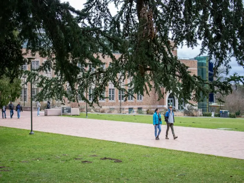 Students walking along a sunny campus path past a brick building